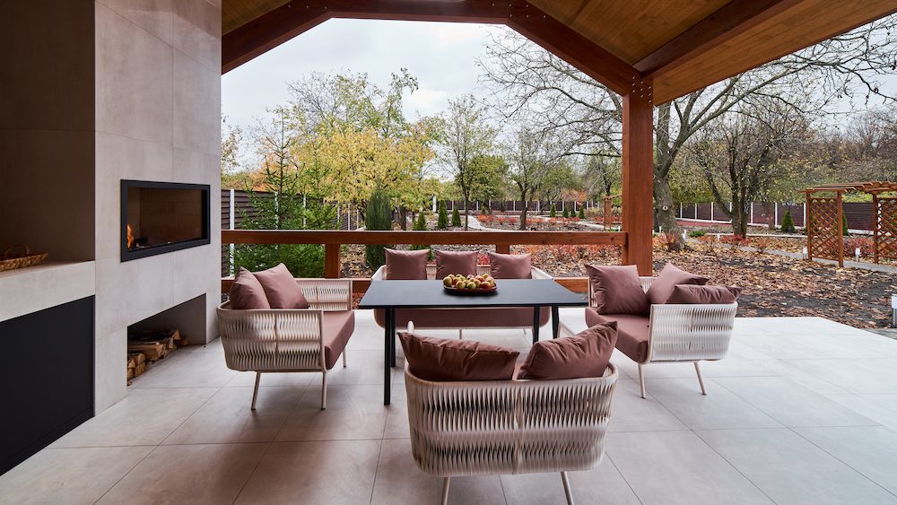 Covered outdoor patio with a modern built-in fireplace, cozy seating, and a view of a serene backyard with trees and fall foliage.