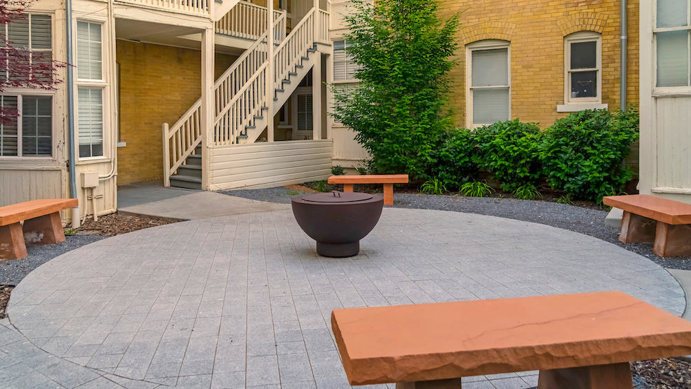 Panorama frame Fire pit on circular pavement with stone benches against building exterior. Residential landscape with buildings featuring outdoor stairs, and white and stone brick walls. Patio made of pavers.
