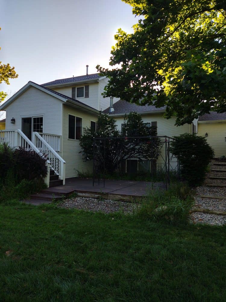 Before photo of project done by Torchwood Landscaping: A New Deck and Patio in Ada, MI. Image shows the back deck and backyard with overgrowth of plants.