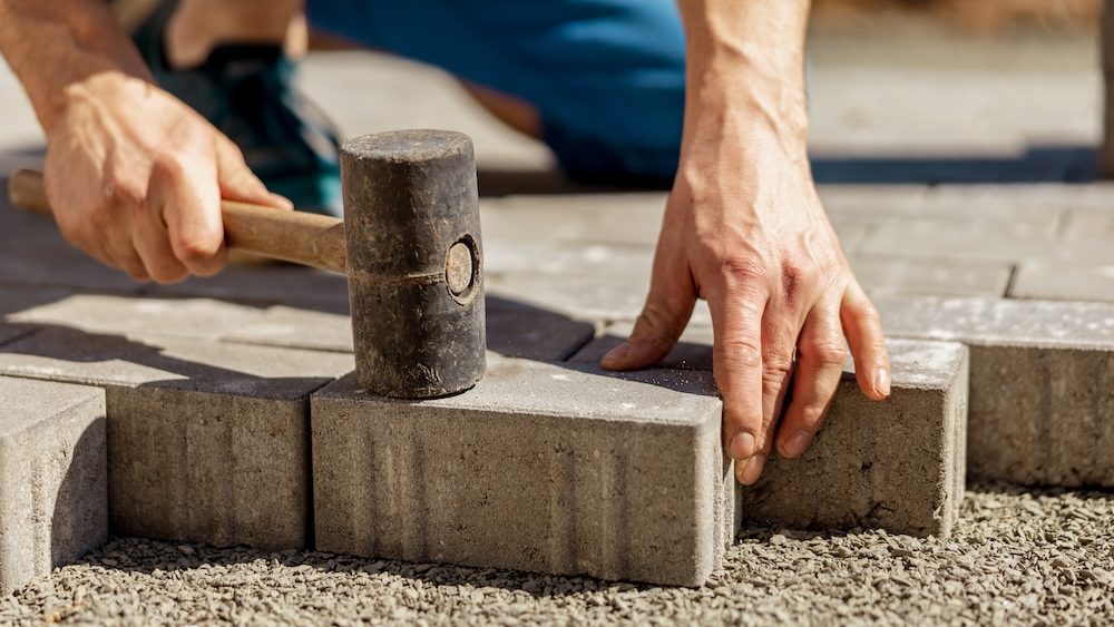 Young man laying gray concrete paving slabs in house courtyard on gravel foundation base. Master lays paving stones. Garden brick pathway paving by professional paver worker. Repairing sidewalk