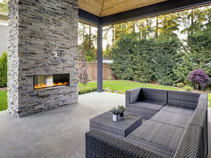 Concrete patio. New modern home features a backyard with covered patio accented with stone fireplace, vaulted ceiling with skylights and furnished with gray wicker sofa placed on concrete floor. Northwest, USA